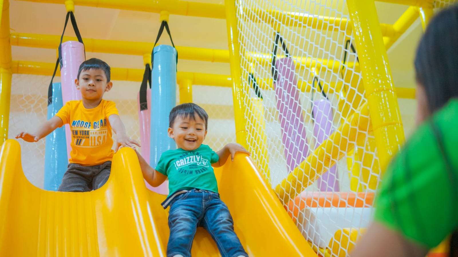Dos niños pequeños sonrientes bajan por un tobogán amarillo en un área de juegos techada